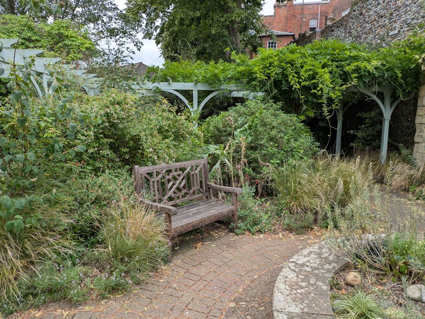 A small outdoor garden corner featuring a weathered wooden bistro table with a matching chair, both with slatted surfaces and metal frames. The chair has a padded cushion with a floral pattern tied to